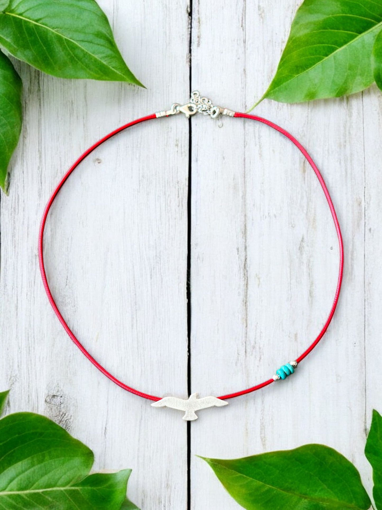 Silver seagull pendant on red cord necklace, elegantly displayed with green leaves on wooden background.