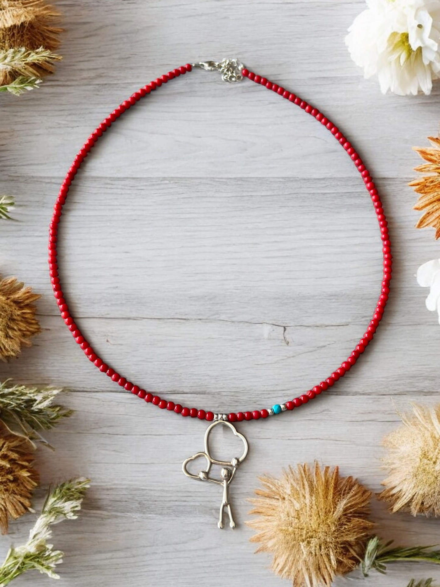 Two Hearts Coral Gemstone Necklace with a silver pendant showing two hearts held by a figure, placed on rustic wooden background with flowers